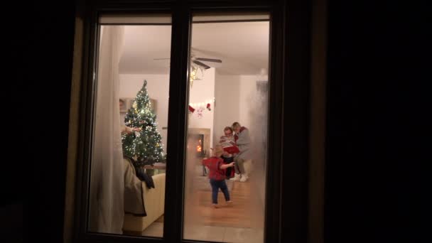 Vista a través de la ventana de la casa helada de la familia disfrutando de la celebración de Navidad cerca de la chimenea. Familia feliz celebrando la Navidad — Vídeos de Stock