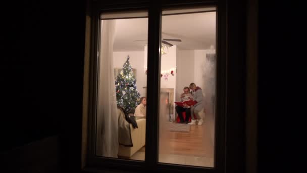 Happy family celebrating christmas. View through frost house window of family enjoying Christmas celebration near the fireplace — Stock Video