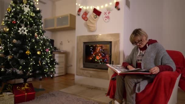 Donna anziana nonna rilassarsi su una poltrona di fronte alla famiglia albero di Natale libro di lettura. Camino. Natale, Capodanno interno — Video Stock