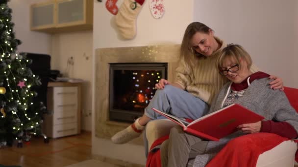 Η ενήλικη κόρη και η μητέρα της περνάνε τα Χριστούγεννα μαζί. Γυναίκες που κοιτάζουν ένα φωτογραφικό άλμπουμ ενώ κάθονται σε μια πολυθρόνα κοντά στο τζάκι και σε ένα χριστουγεννιάτικο δέντρο — Αρχείο Βίντεο