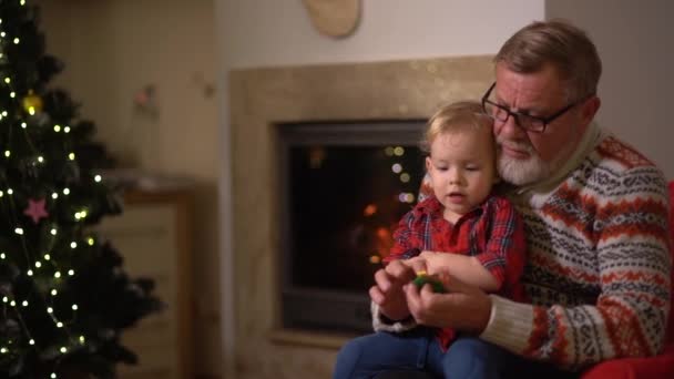 Farfar och barnbarn sitter i en fåtölj nära brasan och en julgran. Familjetraditioner, äldre man och barn ha kul på julafton — Stockvideo