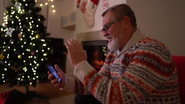 Akıllı telefonlu yaşlı adam ailesini Noel ağacı ve şöminenin yanında bir video görüşmesi yapıyor. Karantina sırasında noel zamanı covid-19 koronavirüs. Yalnız Noel, yeni normal