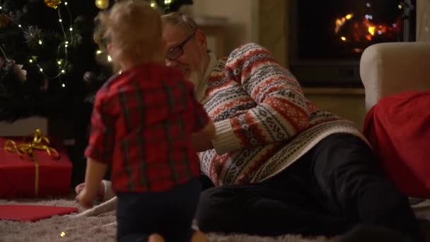 Ältere Männer und Kinder feiern Weihnachten, eine glückliche Familie. Großvater und Enkel und der Hund haben Spaß auf dem Teppich neben dem Weihnachtsbaum liegend. Familientraditionen — Stockvideo