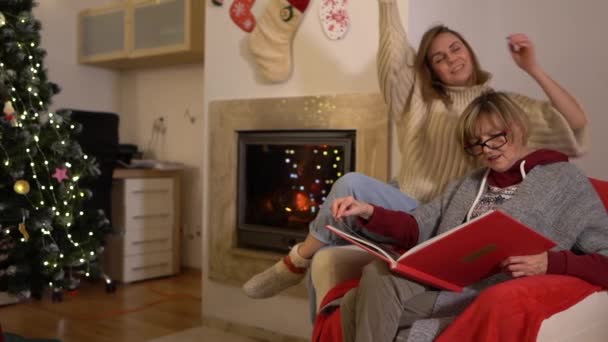 Kvinnor tittar på ett fotoalbum när de sitter i en fåtölj nära eldstaden och en julgran. Vuxen dotter och hennes mamma tillbringar julafton tillsammans — Stockvideo