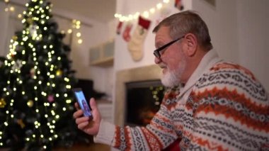 Karantina sırasında noel zamanı covid-19 koronavirüs. Yalnız Noel, yeni normal. Akıllı telefonlu yaşlı adam ailesini Noel ağacı ve şöminenin yanında bir video görüşmesi yapıyor.