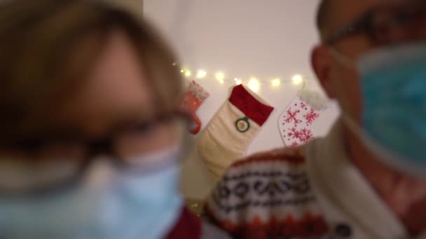 Una pareja madura conoce a la pandemia del covid-19 navideño. Marido y esposa con máscaras médicas protectoras en una casa con decoraciones navideñas y una chimenea. Nuevo concepto normal de vacaciones de Navidad — Vídeos de Stock