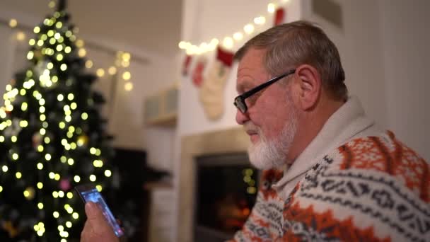 De oude man met een smartphone maakt een videogesprek in de buurt van de kerstboom — Stockvideo