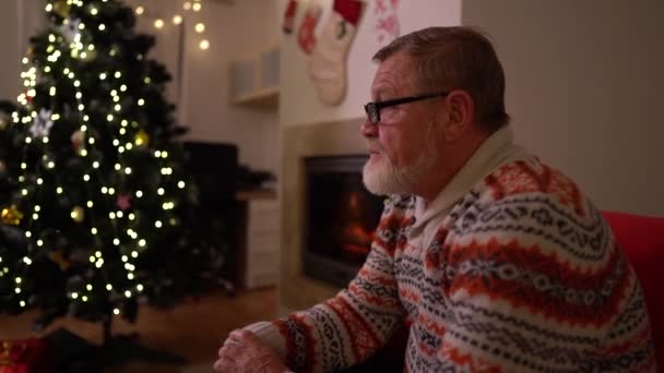 Um homem idoso de barba cinza com óculos e uma camisola de malha senta-se sozinho junto à lareira e a uma árvore de Natal. A celebrar o Natal sozinho. Lar de idosos, quarentena e natal — Vídeo de Stock