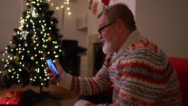 El anciano con un smartphone hace una videollamada cerca del árbol de navidad — Vídeos de Stock