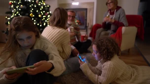 Noel arifesinde Noel ailesi şöminenin yanında toplandı. Yaşlı ebeveynler, yetişkin bir kızı ve iki genç torunu var. Gençler akıllı telefonlarıyla meşgulken yetişkinler çay içiyor. — Stok video