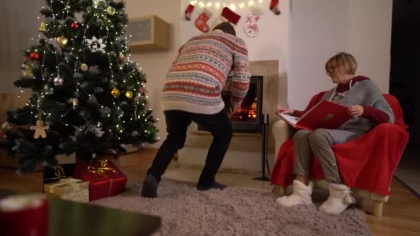 Äldre par sitter nära öppen spis dekorerad till jul. Mysigt hem. Mannen tänder eldstaden, frun sitter i en bekväm fåtölj. Mogna par firar julafton — Stockvideo