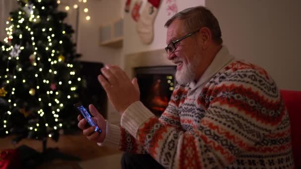 O velho com um smartphone fazer uma chamada de vídeo perto da árvore de Natal — Vídeo de Stock