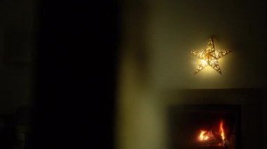 Ağaçtaki Noel ışıkları. Şöminede canlı bir ateş yanıyor. Şöminenin üzerinde bir Noel yıldızı asılı. Noel için dekore edilmiş oturma odası.