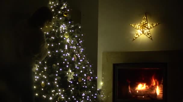 Uma mulher decora uma árvore de Natal perto de uma lareira em chamas. Véspera de Natal. Aconchegante sala de estar decorada para o Natal — Vídeo de Stock