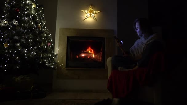 En ung kvinde sidder alene ved pejsen og et juletræ med en telefon i hænderne. Social afstand. Jul under lockdown, nytår og den covid-19 coronavirus pandemi – Stock-video