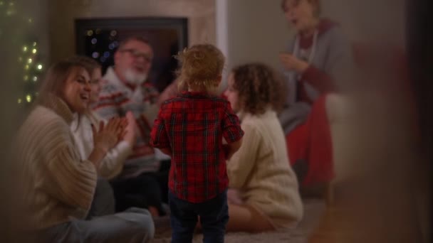 Szczęśliwa rodzina, starsza para, ich córka, wnuczek i pies bawią się przy kominku w świąteczny wieczór. Rodzina świętuje Boże Narodzenie, widok przez okno z mrozem — Wideo stockowe