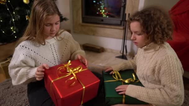 Dwie nastoletnie koleżanki otwierają prezenty od Mikołaja siedząc obok choinki i kominka w przytulnym salonie. Szczęśliwego dzieciństwa, Wigilii — Wideo stockowe