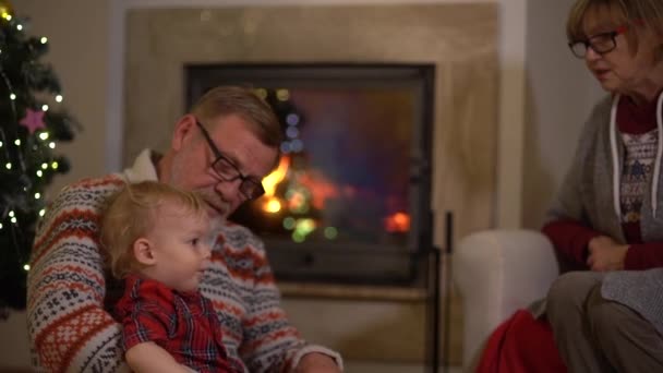 Los abuelos se sientan junto a la chimenea con su nieto en la sala de estar decorada para Navidad. Nochebuena, infancia feliz — Vídeos de Stock