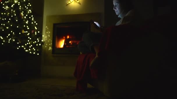 Distance sociale. Une jeune femme s'assoit seule près de la cheminée et d'un sapin de Noël avec un téléphone à la main. Noël pendant le confinement, le nouvel an et la pandémie de coronavirus covid-19 — Video