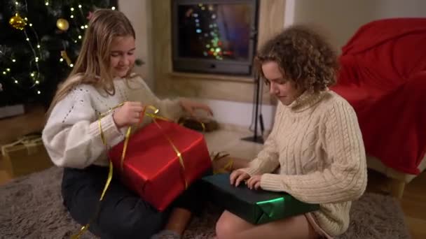 两个少女朋友坐在圣诞树旁和一个舒适的客厅里的壁炉旁，打开圣诞老人送给她们的礼物。快乐的童年，平安夜 — 图库视频影像