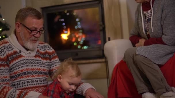 Οι παππούδες κάθονται δίπλα στο τζάκι με τον εγγονό τους στο σαλόνι διακοσμημένο για τα Χριστούγεννα. Παραμονή Χριστουγέννων, ευτυχισμένη παιδική ηλικία. — Αρχείο Βίντεο