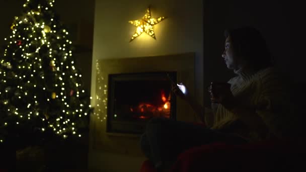 Distância social. Uma jovem senta-se sozinha perto da lareira e de uma árvore de Natal com um telefone nas mãos. Natal durante o confinamento, ano novo e a pandemia do coronavírus covid-19 — Vídeo de Stock