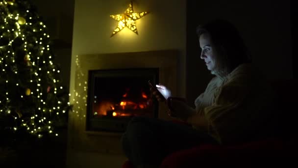 Latar belakang Tahun Baru. Seorang gadis kesepian duduk di dekat perapian di ruang tamu yang didekorasi untuk Natal. Lampu di pohon Natal. Normalitas baru, pengurungan coronavirus covid-19. Latar belakang Tahun Baru — Stok Video
