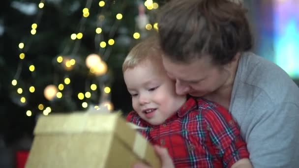 Μητέρα και παιδί κοντά στο χριστουγεννιάτικο δέντρο. Ένα μικρό αγοράκι με τη μαμά του να κοιτάει με ενθουσιασμό μέσα στο κουτί των Χριστουγέννων. Ευτυχισμένη παιδική ηλικία, γιορτάζει τα Χριστούγεννα και το νέο έτος στο σπίτι — Αρχείο Βίντεο