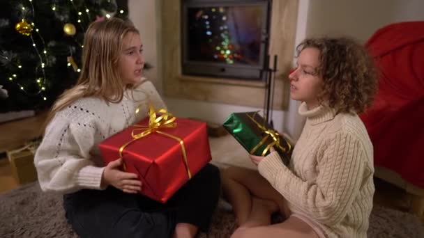 Boldog gyermekkort, Szenteste. Két tizenéves lány barátok kinyit ajándékokat a Mikulás, miközben ül közel egy karácsonyfa és kandalló egy hangulatos nappaliban — Stock videók