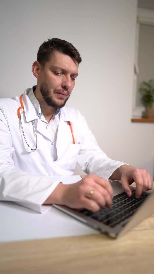Dikey video. Bir adam otolaryngologist bir hastaya çevrimiçi danışır ve bir dizüstü bilgisayar kullanarak veri kaydeder. Salgın sırasında ilaç, teletıp konsepti — Stok video