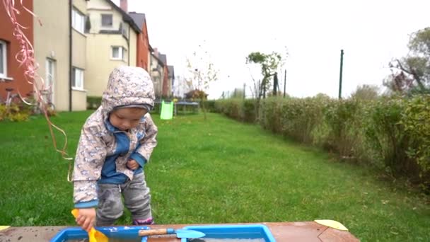 Un niño de dos años juega con una cucharada amarilla en una caja de arena al aire libre en un jardín de infantes. El niño está vestido con botas y una chaqueta de otoño. Actividad infantil — Vídeos de Stock