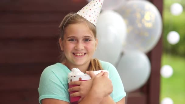 Ragazza adolescente felice in un cappello di compleanno tiene un regalo in mano. Ritratto della ragazza di compleanno, compleanno dei bambini, concetto di ristorazione — Video Stock