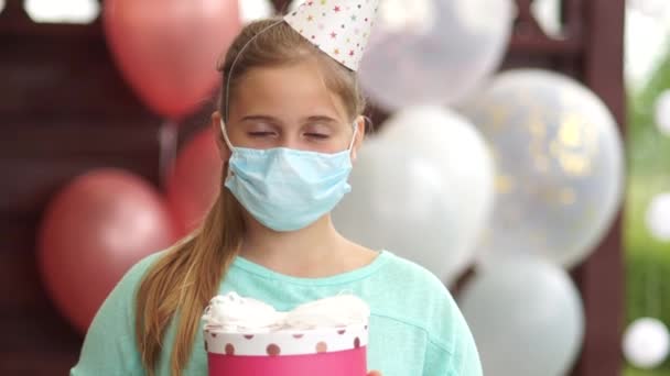 Triste petite fille avec une boîte cadeau dans ses mains à sa fête d'anniversaire porter un masque pour prévenir le virus. Fête de fond coloré avec des ballons. Anniversaire seul. Restez à la maison en quarantaine — Video