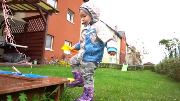 Маленький мальчик в сапогах и куртке играет с песочницей рядом с домом. Детский досуг, счастливое детство — стоковое видео