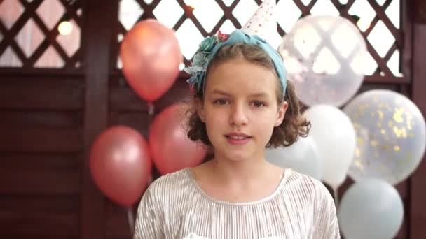Közelkép egy szülinapos lányról a lufik, a gyerekek születésnapja, a vendéglátás hátterében. Boldog tinédzser lány szülinapi kalapban egy ajándékot tart a kezében. — Stock videók