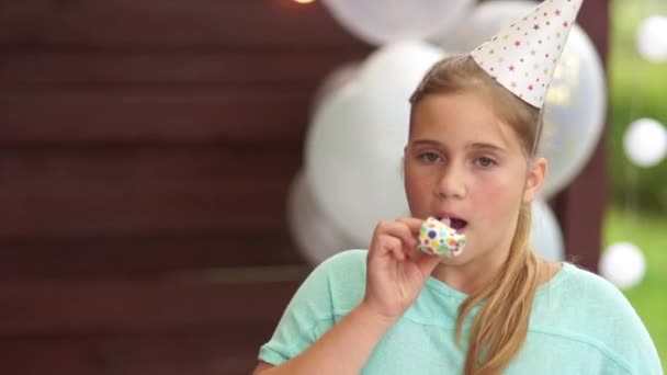 Una adolescente con un sombrero de cumpleaños sopla una pipa y se ríe. Feliz infancia, fiesta de cumpleaños de los niños al aire libre — Vídeo de stock