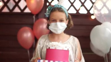 Maskeli güzel bir doğum günü kızının yakın portresi balonların arka planına karşı bir hediye tutuyor. Karantina sırasında yalnız doğum günü Coronavirus covid-19, yeni normal
