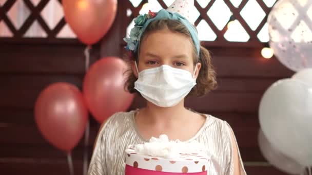 Magányos születésnap a karantén alatt Coronavirus covid-19, az új normális. Egy gyönyörű, maszkos szülinapos lány portréja, aki ajándékot tart az ünnepi lufik hátterében.