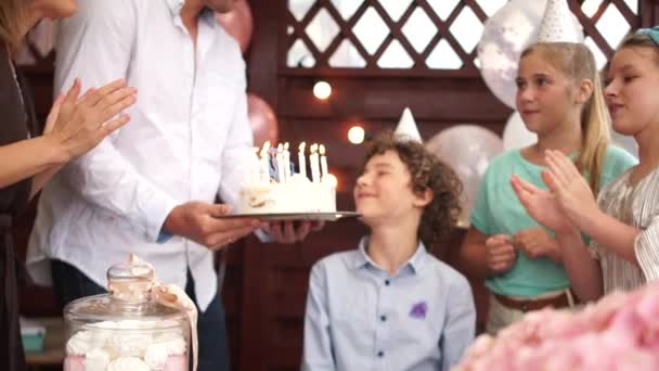 生日快乐的男孩正在他的家人和朋友的簇拥着下着的生日蛋糕上吹灭蜡烛。一个友善的家庭在院子里庆祝他们十几岁儿子的生日 — 图库视频影像