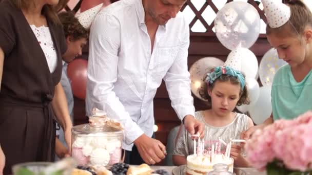 La cumpleañera, los padres y los invitados encienden velas en el pastel de cumpleaños. Cumpleaños de niños. Celebración familiar o una fiesta en el jardín en el patio trasero — Vídeos de Stock