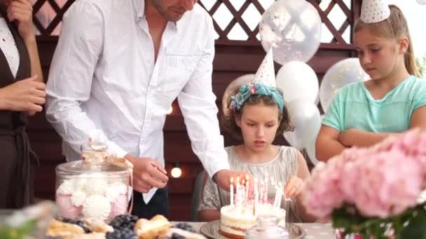 Narozeninová dívka, rodiče a hosté zapalují svíčky na narozeninovém dortu. Děti mají narozeniny. Oslavenkyně se zašklebí, když čeká na svíčky na dortu. Přej si něco — Stock video
