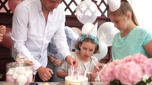 裏庭の外で家族のお祝いやガーデンパーティー。誕生日の女の子、両親、ゲストは誕生日ケーキにろうそくを灯します。子供の誕生日 — ストック動画