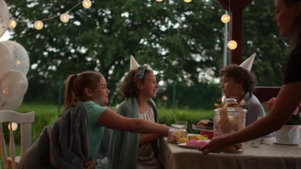 Kinderverjaardag buiten, catering concept. Vrolijke kinderen die bessen eten terwijl ze aan de feestelijke tafel zitten — Stockvideo