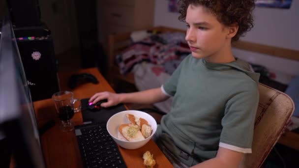 Un colegial juega un juego de computadora y come sentado en una mesa en su habitación. Adicción informática entre adolescentes — Vídeo de stock