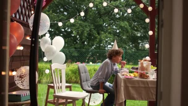 Triste garçon sur son anniversaire est assis à une table sur laquelle il ya des friandises et un gâteau avec l'inscription Joyeux anniversaire. Anniversaire solitaire pendant la pandémie de coronavirus covid-19 — Video