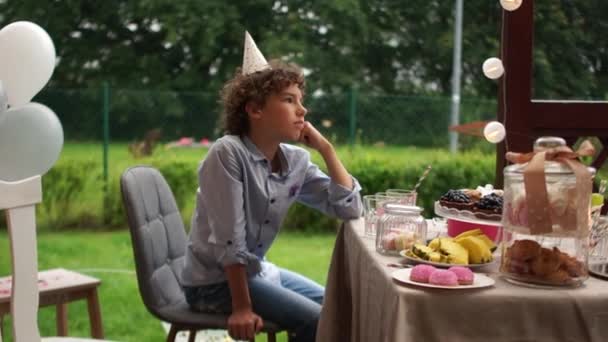 Gros plan portrait d'un adolescent triste et solitaire assis à la table d'anniversaire. Un garçon aux cheveux bouclés dans un chapeau d'anniversaire passe son anniversaire sans amis — Video