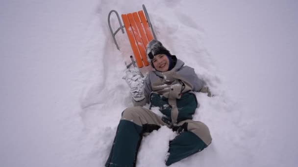 Adolescent garçon est tombé dans la neige d'un traîneau et agite sa main. Concept d'activités hivernales, de loisirs et de divertissement pour enfants — Video