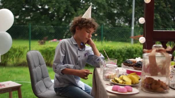 Un adolescent triste dans un chapeau d'anniversaire s'assoit à une table d'anniversaire servie. Un anniversaire solitaire sans amis lors d'un confinement COVID-19 — Video