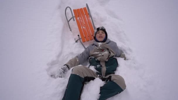 Ευτυχισμένο σχολιαρόπαιδο βρίσκεται στο χιόνι κατά τη διάρκεια των χειμερινών διακοπών. Το αγόρι έπεσε από ένα έλκηθρο στο χιόνι, διασκεδαστικές χειμερινές δραστηριότητες, παιδική διασκέδαση — Αρχείο Βίντεο