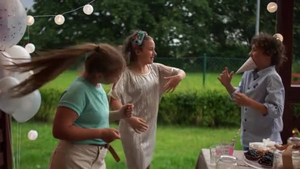 Drei Schulfreunde tanzen auf einer Geburtstagsparty. In der Nähe gibt es eine Geburtstagstafel mit Kuchen und Gebäck. Glückliche Kindheit — Stockvideo